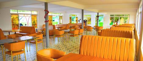A spacious indoor dining area with orange seating and tables, large windows with greenery visible outside, and a tiled floor.