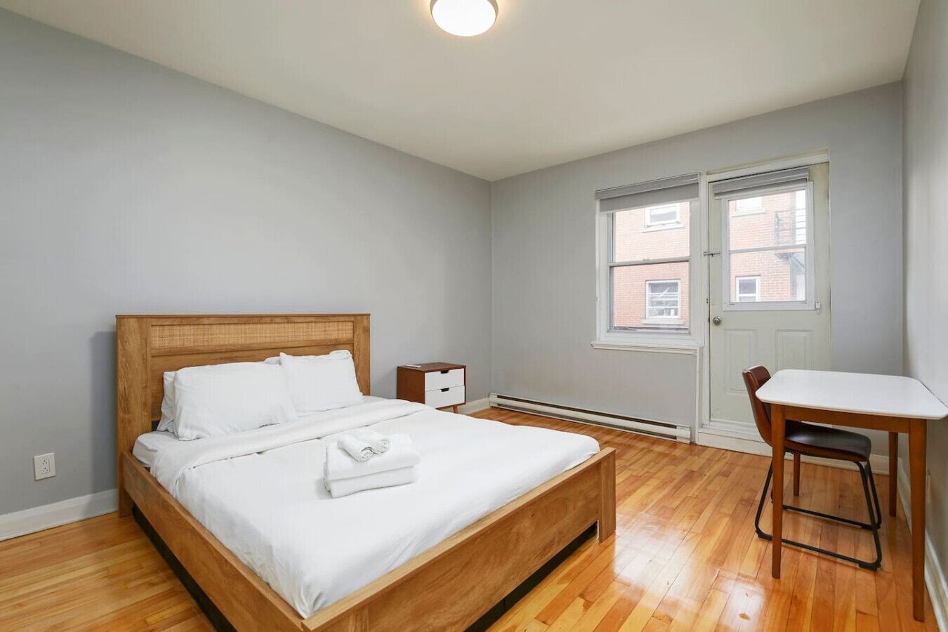 Quarto com cama de madeira, mesa de cabeceira, escrivaninha e janela com vista para um prédio de tijolos.