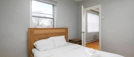 A bedroom with a wooden headboard, a single bed with white bedding, a small bedside table, and a window with blinds.