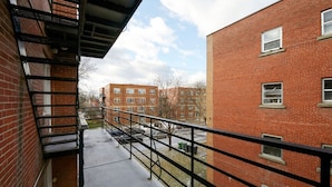 Ein Balkon mit Metallgeländer, der auf einen Parkplatz und angrenzende Backsteingebäude blickt.