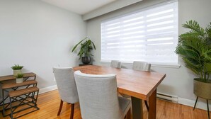 A modern dining area with a wooden table and grey upholstered chairs, a potted plant, and a large window with blinds.