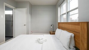 Ein Schlafzimmer mit einem Bett, einem Nachttisch, einer Lampe, einer Tür zu einer Küche und einem Fenster.