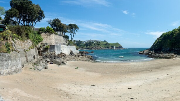 Uma praia com um muro de pedra, árvores e vista para o mar.