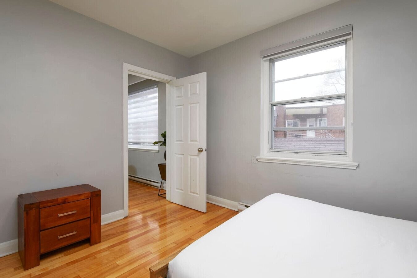 A bedroom with a wooden dresser, a bed, a window with blinds, and a door leading to another room.