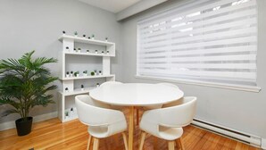 Een moderne eethoek met een ronde tafel en twee witte stoelen, een witte boekenkast met planten, en een groot raam met jaloezieën.