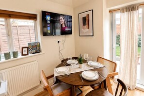 A dining area with a round table set for four, a television mounted on the wall, and a window with blinds.