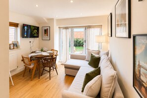 Una sala de estar con sofá, una mesa de comedor con sillas, un televisor montado en la pared y una ventana con cortinas.