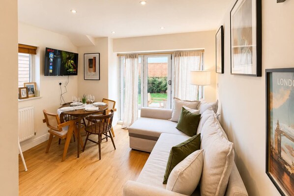 A living room with a sofa, a dining table with chairs, a TV mounted on the wall, and a window with curtains.