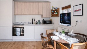 A modern kitchen with white cabinets, a dining area with wooden chairs and a table set for four, a microwave, and a television on the wall. - Queens Park Apt w/ mini garden near Notting Hill (London)