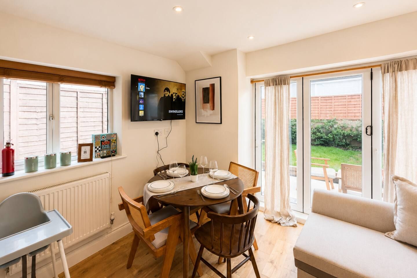 A dining area with a table set for four, a television mounted on the wall, and a sofa with a view to the outdoors.