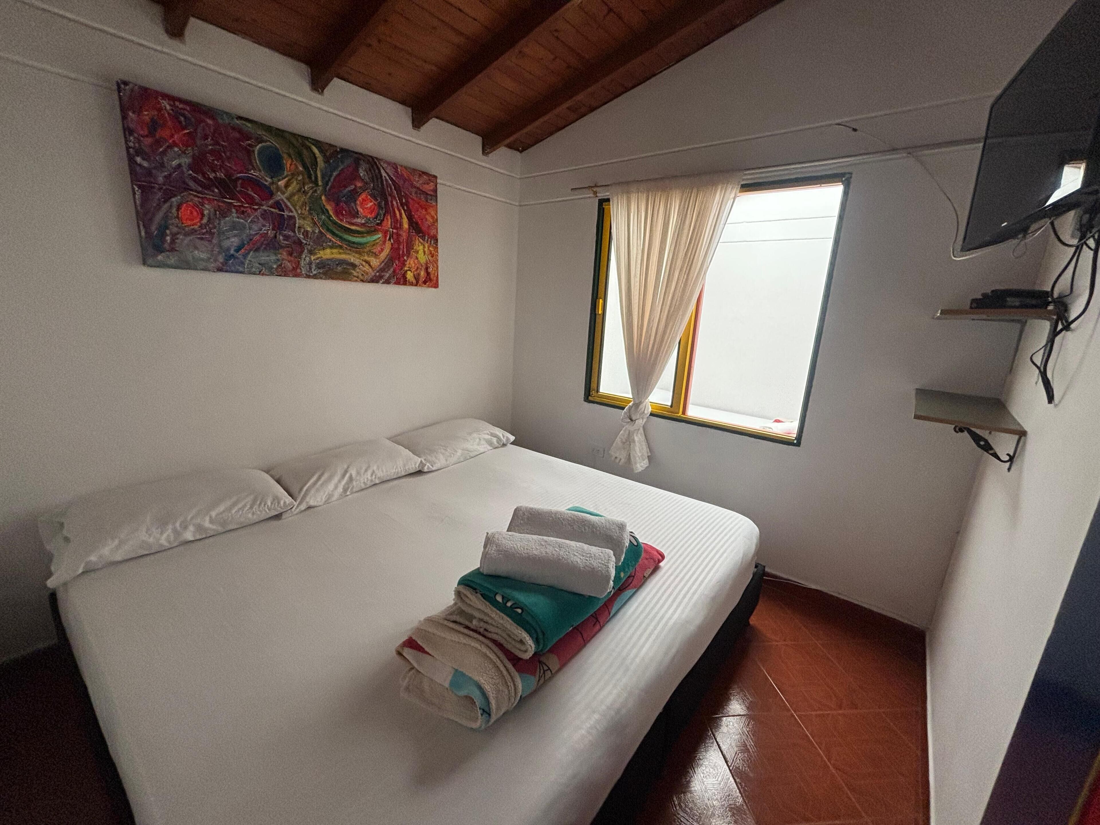 Un dormitorio con cama, almohadas, un cuadro abstracto de colores, una ventana con cortinas y un televisor montado en la pared.