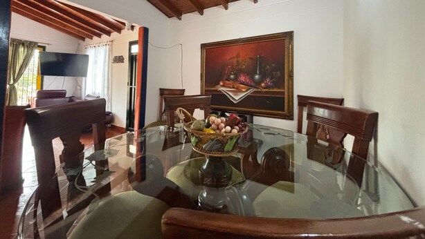 A dining area with a glass table, wooden chairs, a painting on the wall, and a television in the background. - Apartamento Zócalos de Guatape (Guatape)