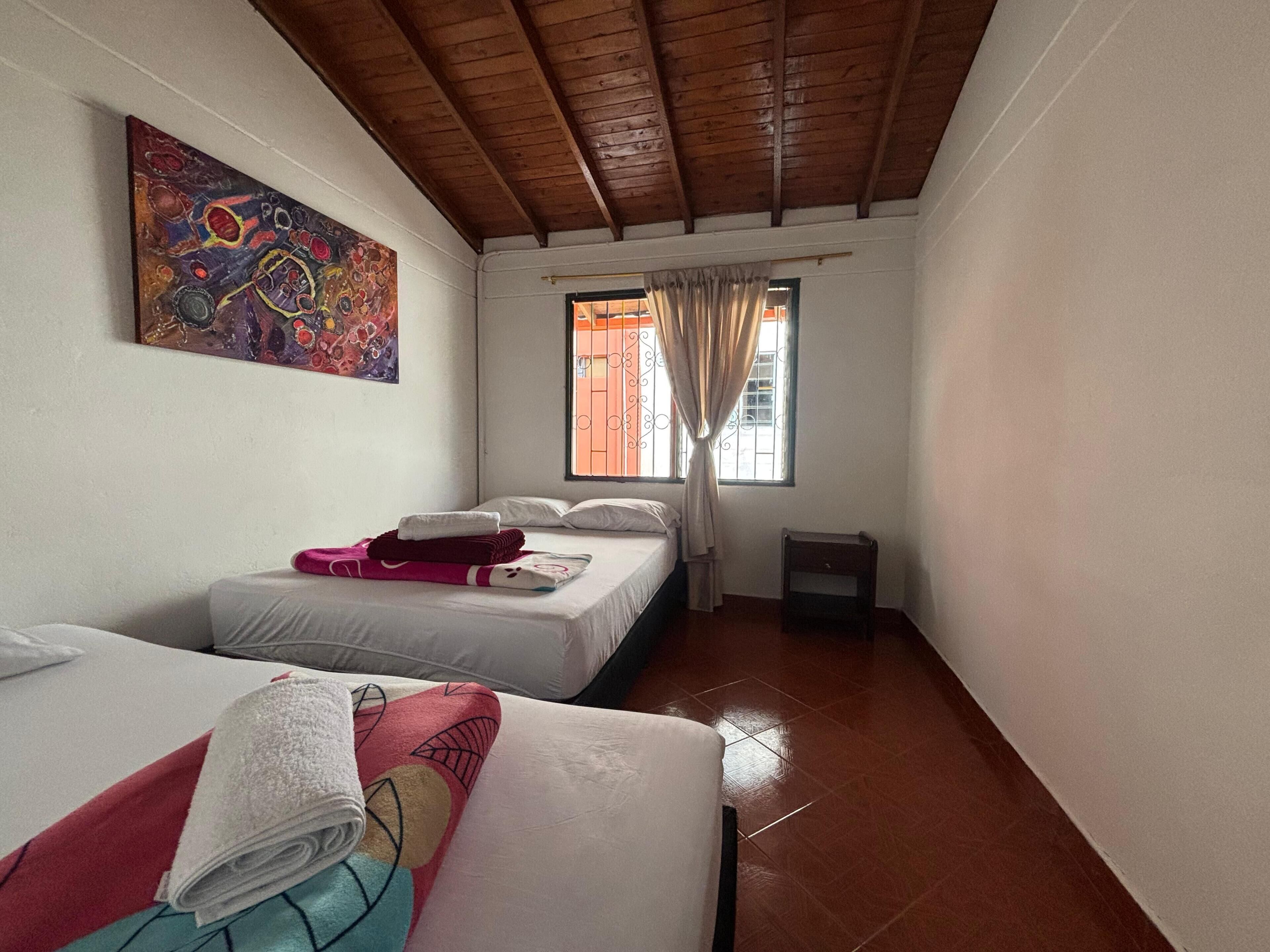 A room with two beds, a window with curtains, a painting on the wall, and a tiled floor.