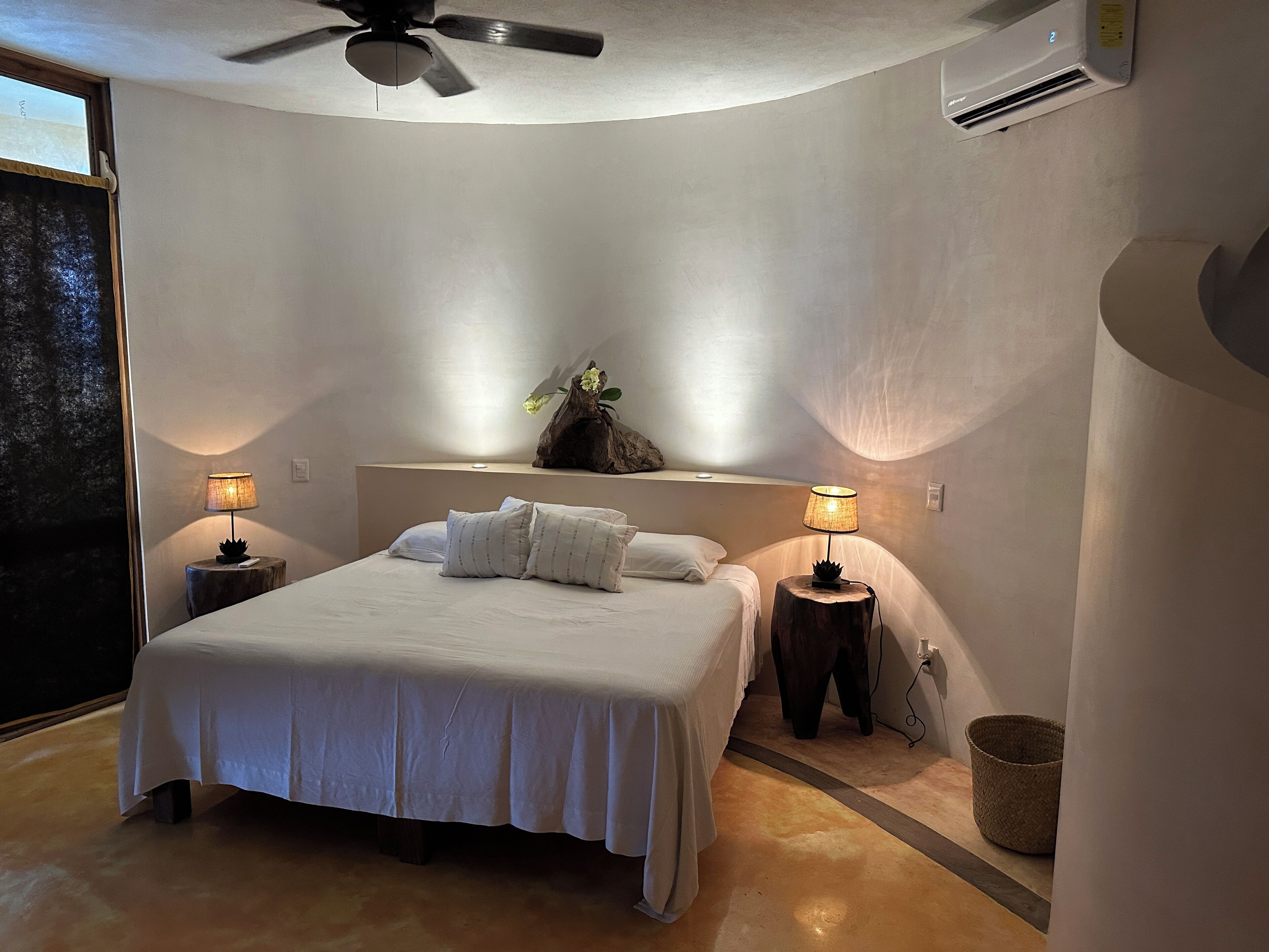 Un dormitorio con una cama, dos mesitas de noche con lámparas, un ventilador de techo y una ventana.