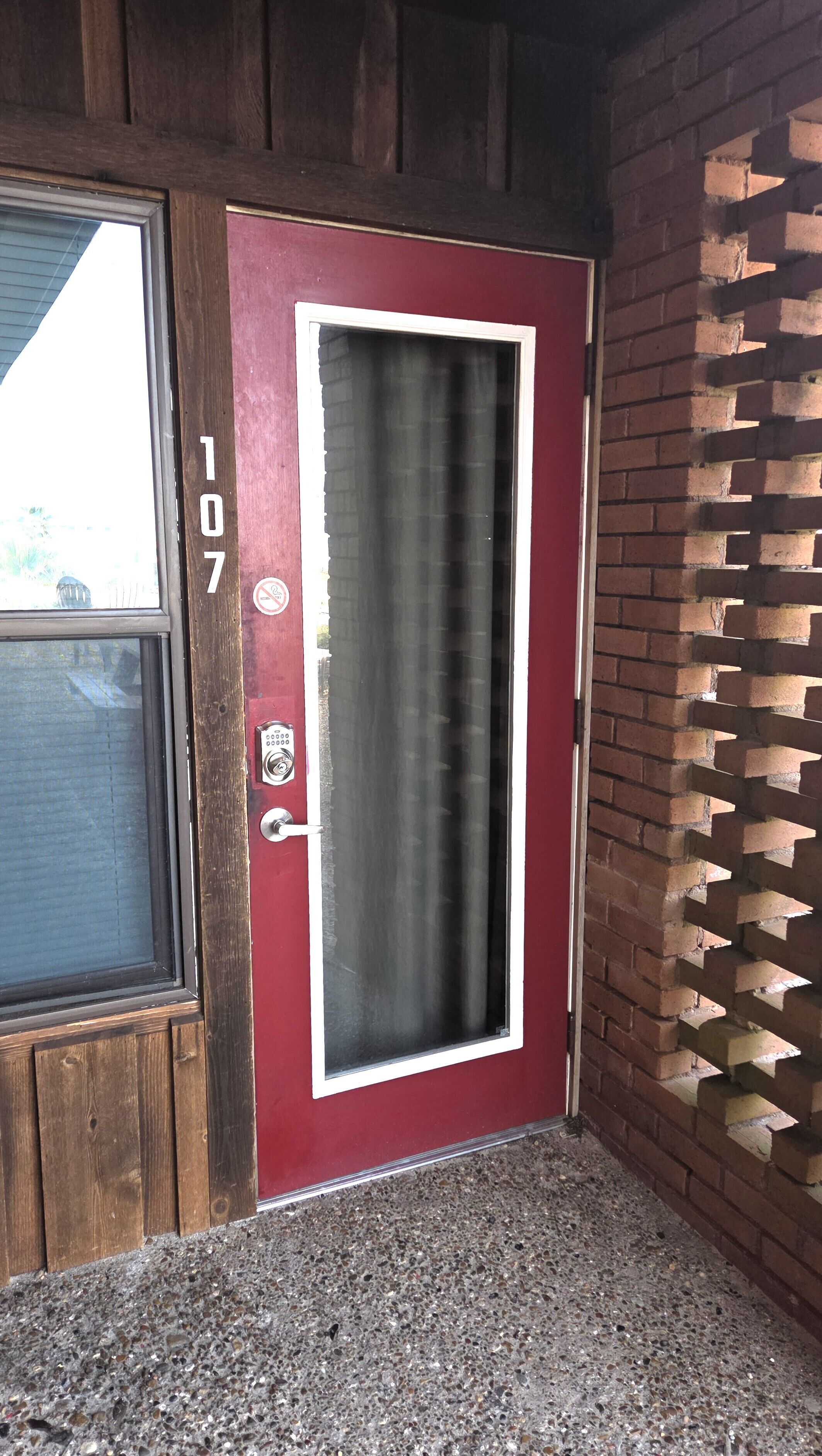 Une porte rouge avec une fenêtre, un système de verrouillage à clavier et un mur de briques agrémenté d’une grille de fenêtre.