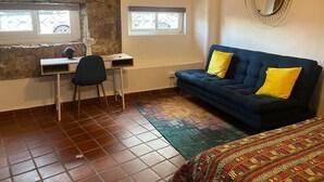 Een kamer met een bureau, stoel, blauwe bank met gele kussens, een kamerkleed met dessin en een spiegel aan de muur.