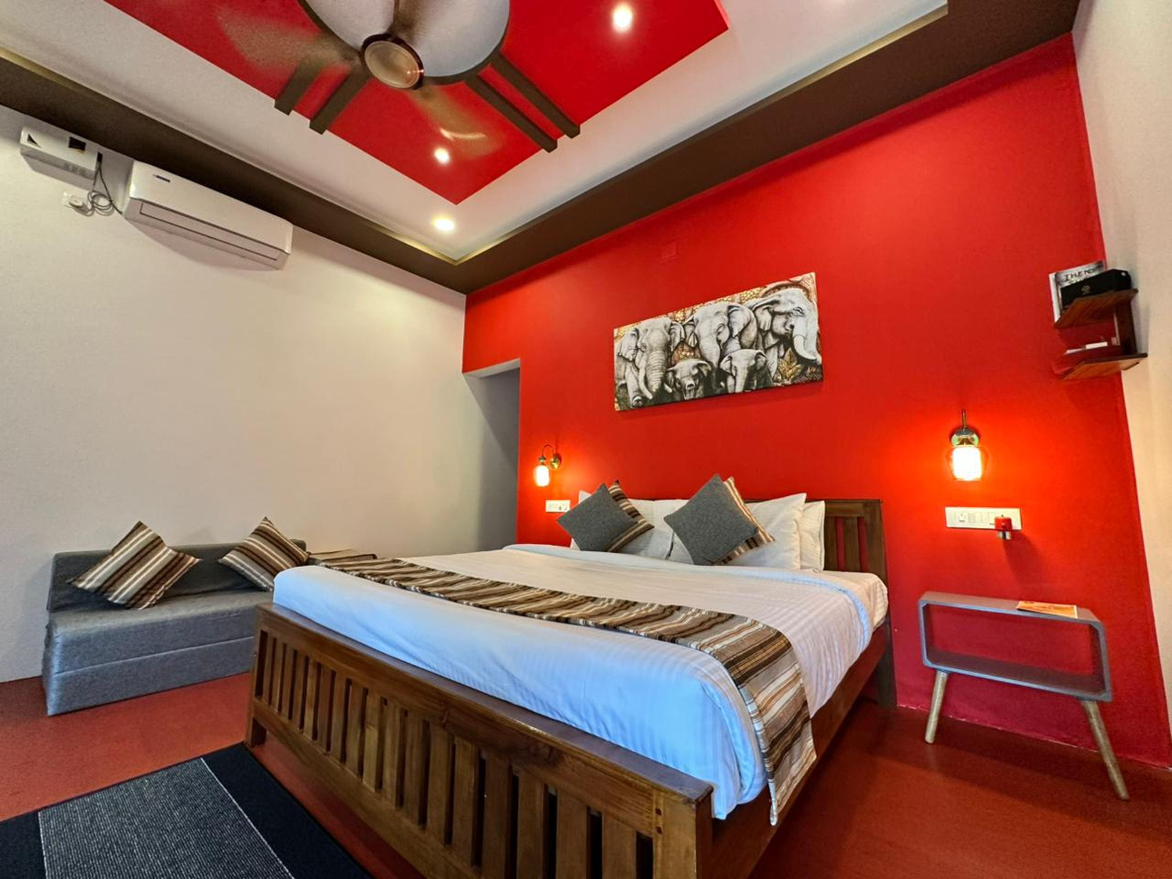 A hotel room with a wooden bed, red walls, a ceiling fan, and a painting of elephants.