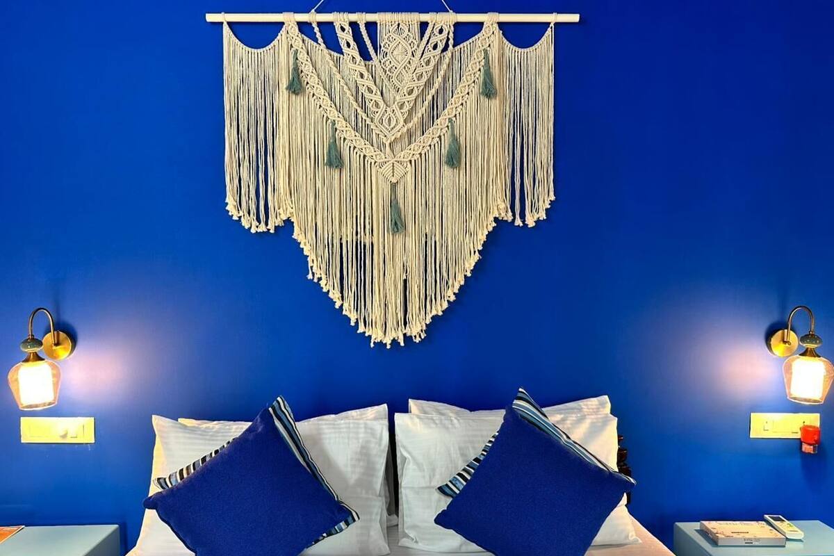 Una habitación de hotel con una pared azul, una cama con ropa de cama blanca y azul, y un tapiz de macramé en la pared.