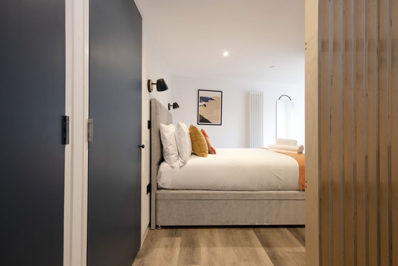 A bedroom with a bed, a wall-mounted lamp, a framed picture, and a window with blinds.