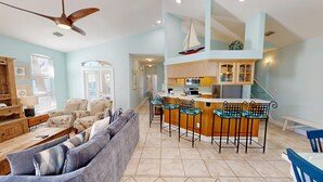 A spacious living room with a large sofa, a wooden coffee table, and a ceiling fan. A kitchen with a bar area and a sailboat decoration is visible in the background. - Mango Tango - Dog Friendly 5 BR home in gated community close to beach (Miramar Beach)