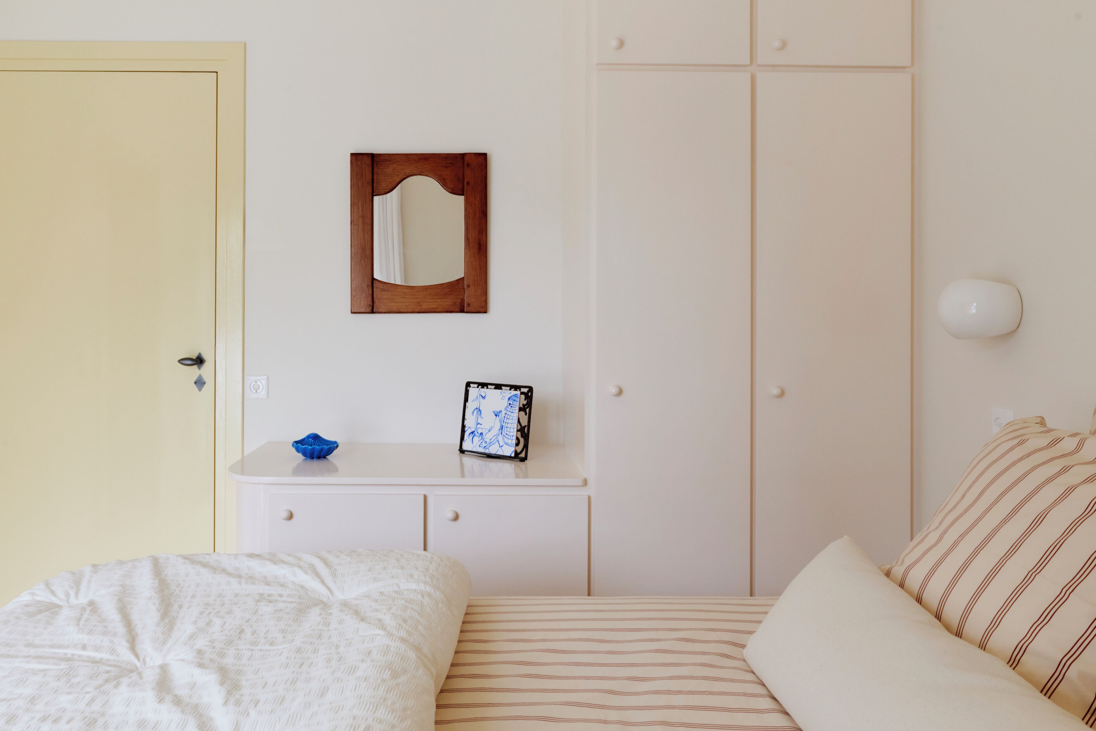 Une chambre à coucher comprenant un lit, une table de chevet avec un miroir et une tablette, une armoire et un luminaire fixé au mur.