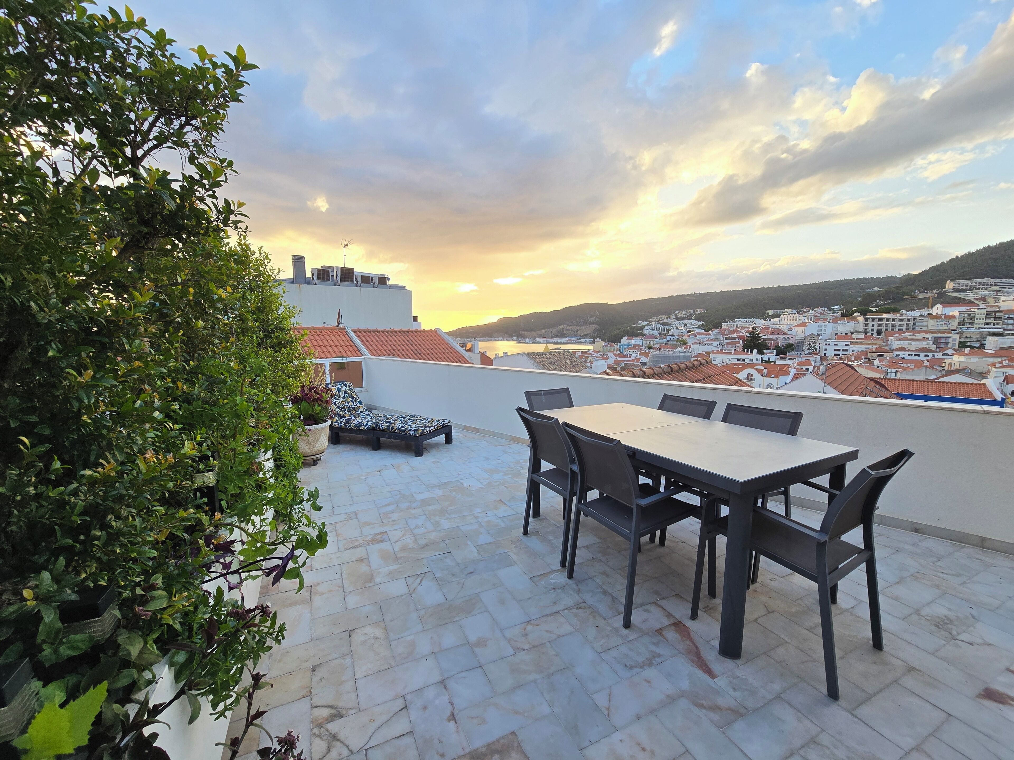 A rooftop terrace with a table and chairs, surrounded by potted plants, overlooking a cityscape with red-roofed buildings.