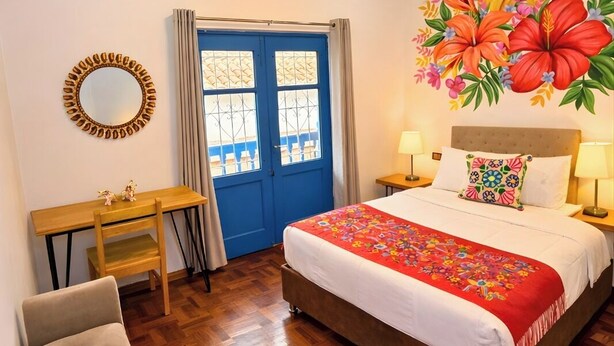 Une chambre d’hôtel avec un lit, un bureau, une chaise, une porte bleue et une fresque murale à motifs floraux.