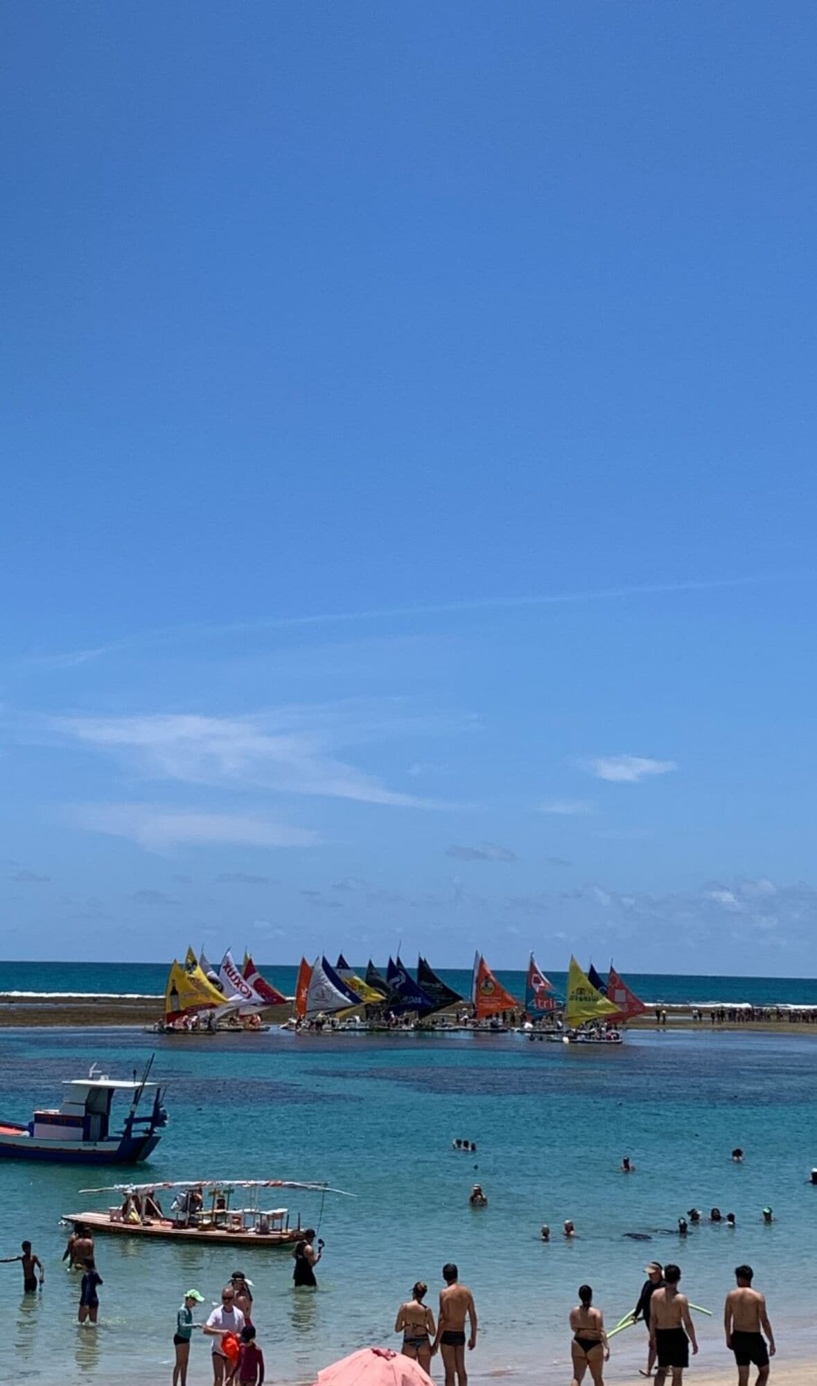 Uma praia com pessoas nadando e caminhando, barcos e bandeiras coloridas na água.