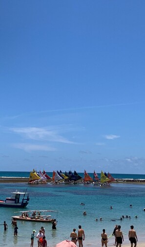 Ein Strand mit badenden und spazierenden Personen, Booten und bunten Flaggen im Wasser.