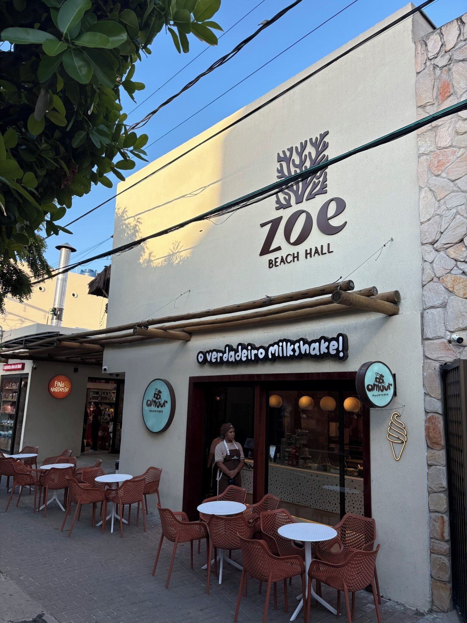 Un café llamado 'Zoe Beach Hall' con mesas al aire libre y un letrero que anuncia 'verdadeira milkshake!'