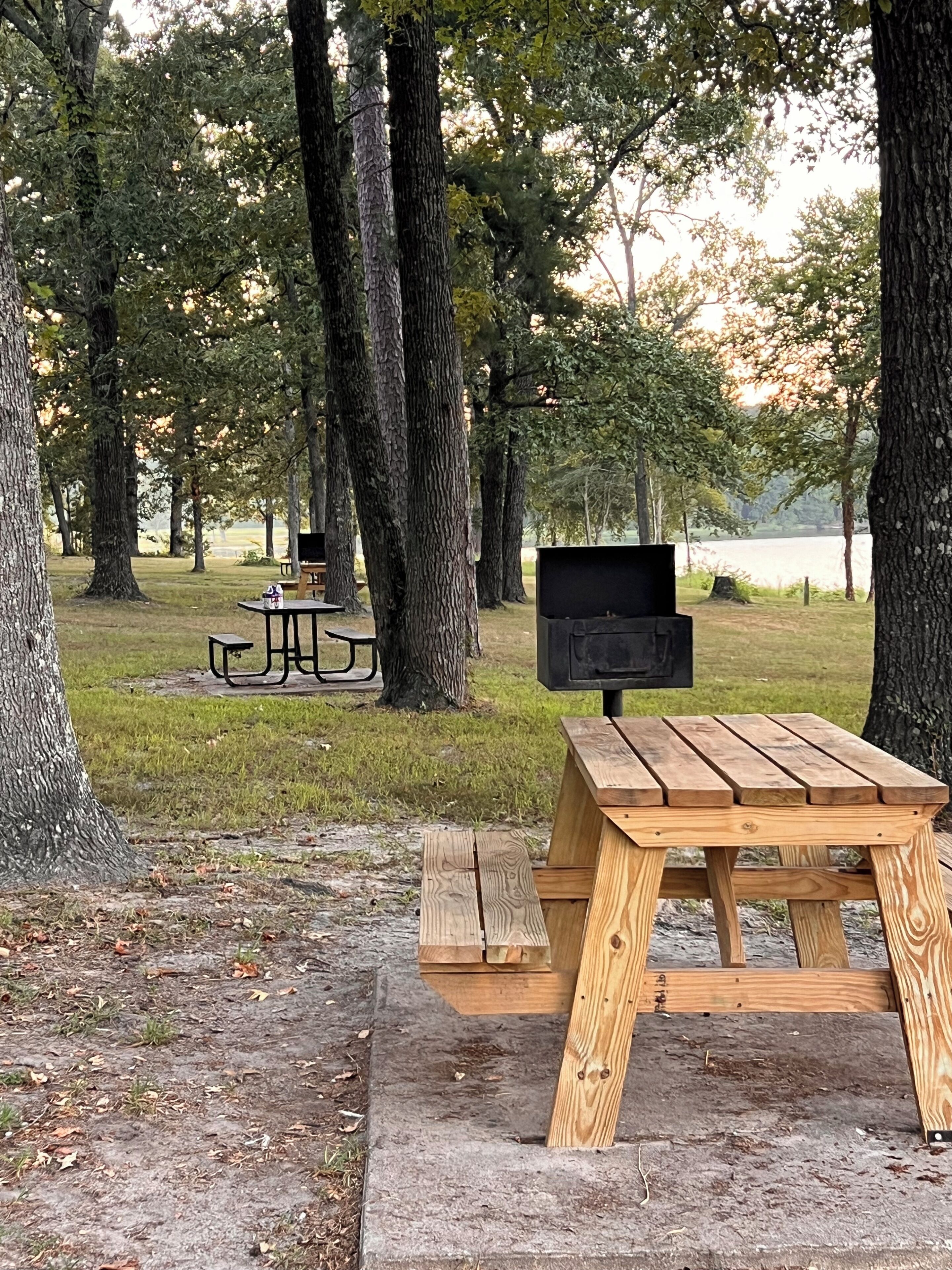 Una mesa de picnic con una parrilla en un parque con árboles y otra mesa de picnic al fondo.