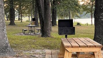 Una mesa de picnic con una parrilla en un parque con árboles y otra mesa de picnic al fondo.