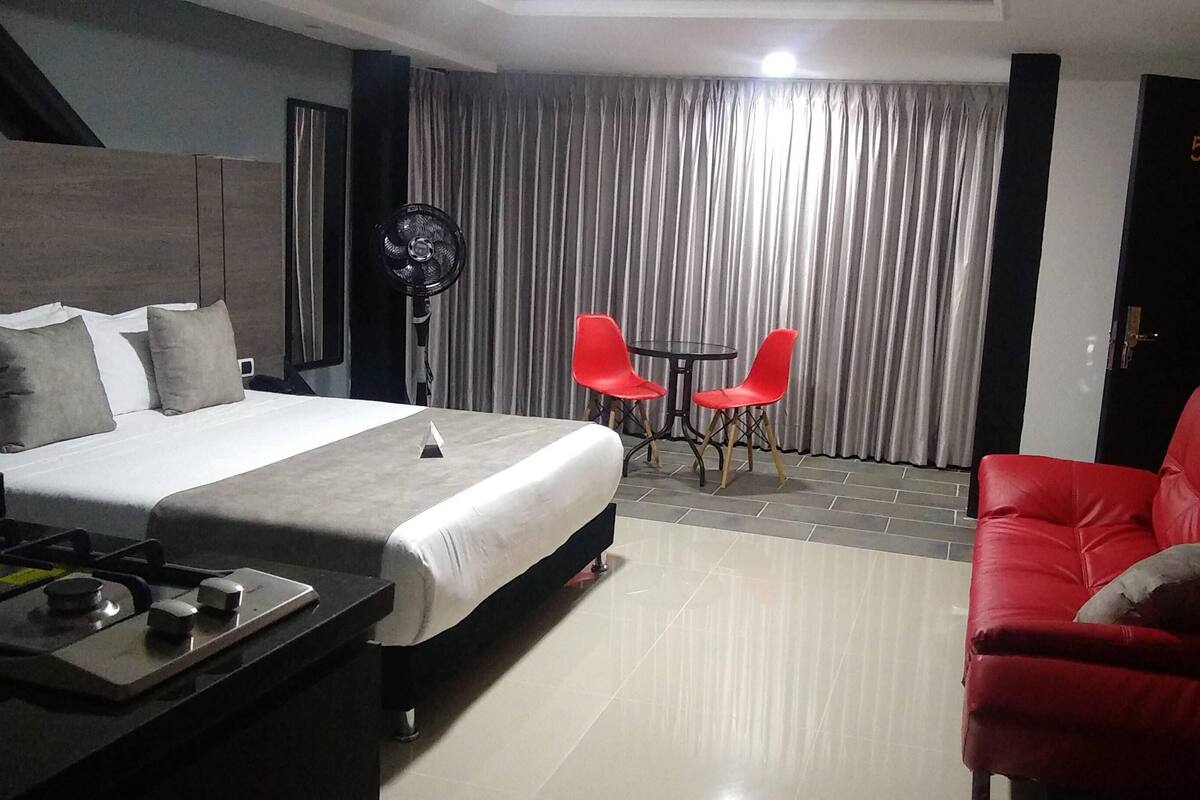 Une chambre d’hôtel avec un lit, un canapé rouge, une petite table et un ventilateur.