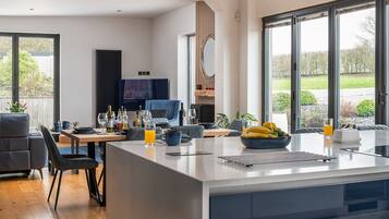 A modern kitchen with a central island, dining area, and large windows offering a view of the outdoors.