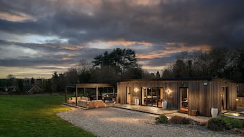 A modern wooden cabin with a patio area, large windows, and a gravel driveway.