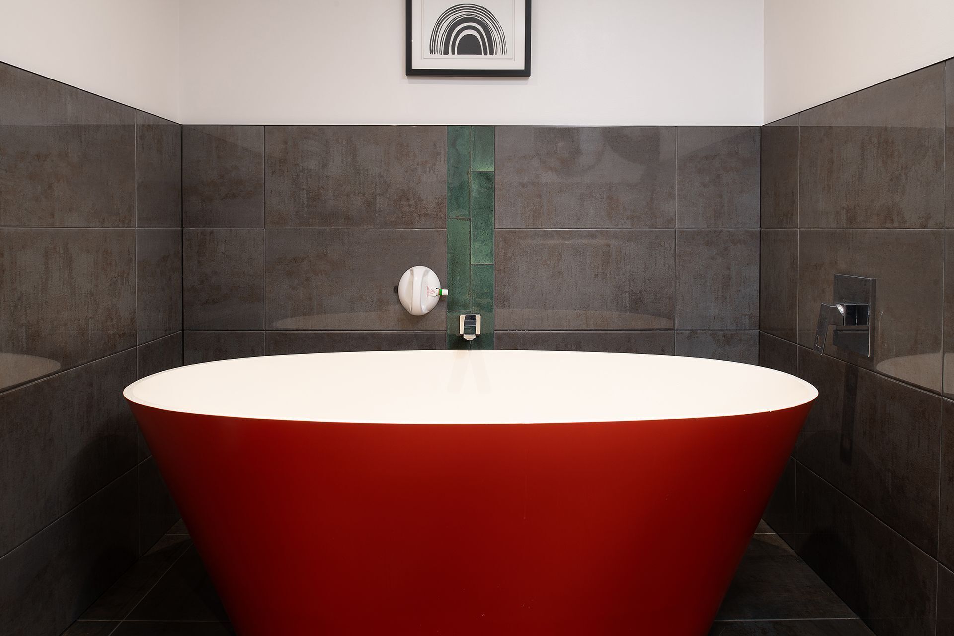 A modern bathroom with a large red bathtub, dark tiled walls, and a single framed artwork on the wall.
