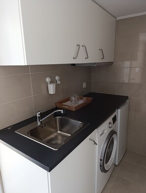 A compact kitchen with a stainless steel sink, a built-in washing machine, and a wooden tray with glasses. - Cascais Studio with kitchenette, bathroom, and access to the terrace (Cascais)