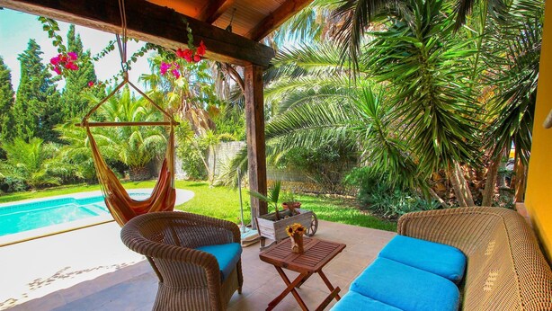 Un espace extérieur aménagé, abrité et doté de mobilier en osier, d’un fauteuil suspendu, et offrant une vue sur une piscine et une végétation luxuriante.