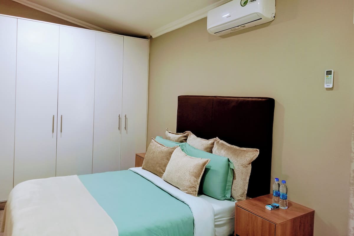 A bedroom with a bed, a wardrobe, an air conditioner, a bedside table with water bottles, and a wall-mounted thermostat.