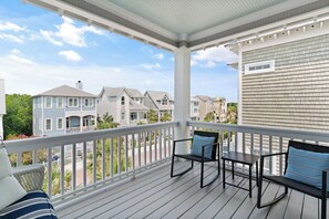A covered porch with a white railing, a black rocking chair, a small table, and a blue cushion. - New Listing! -Satilla Cottage -Coast Cottages (St Simons Island)