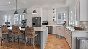 A spacious kitchen with white cabinetry, a central island with bar stools, pendant lighting, and a view of the outside through large windows. - New Listing!  -Satilla Cottage -Coast Cottages (St Simons Island)