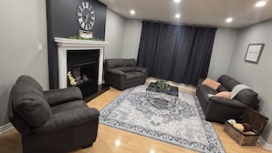 A living room with two dark sofas, a glass coffee table, a patterned rug, a fireplace, and a clock on the wall.