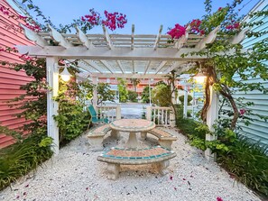 A covered outdoor seating area with a table and benches, surrounded by plants and a pergola. - Perfect Beach Escape - Charming 1 Bedroom Condo Just Steps From The Sand (Bradenton Beach)