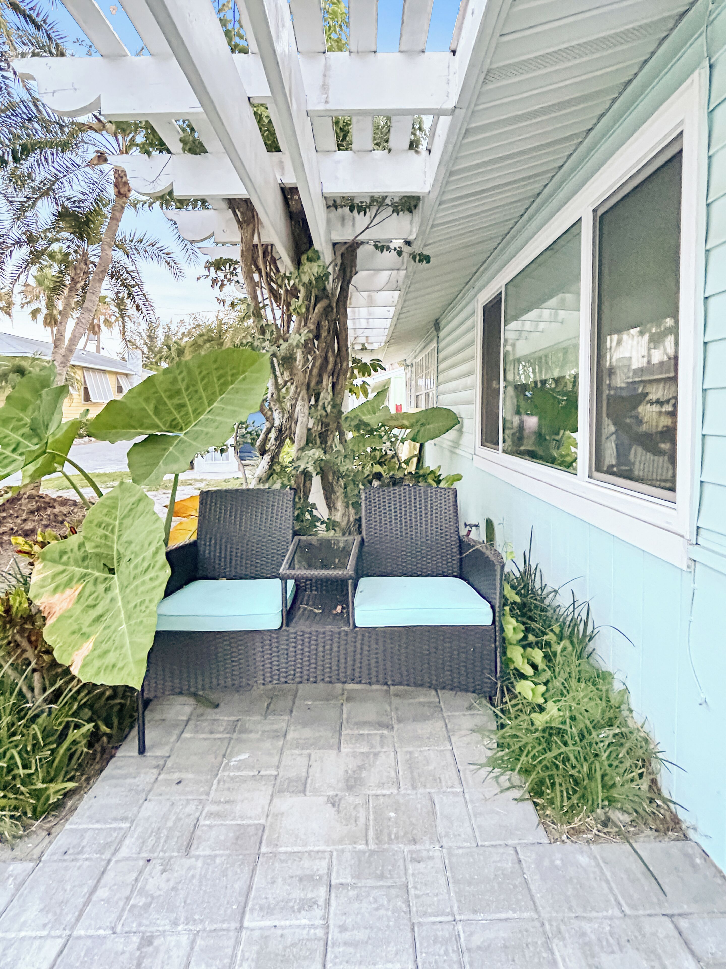An outdoor patio with a wicker sofa and a table, surrounded by large plants and a pergola.