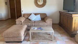 A living room with a beige sofa, a matching ottoman, a coffee table with magazines, a television on a wooden cabinet, and a woven wall hanging.