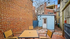 Un mur de briques, un petit abri bleu, une table avec des chaises et une ruelle étroite.