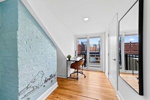 Une pièce avec un plafond en pente, un plancher en bois, un bureau avec une chaise, une fenêtre donnant sur des immeubles et un mur orné d’une fresque représentant des oiseaux.