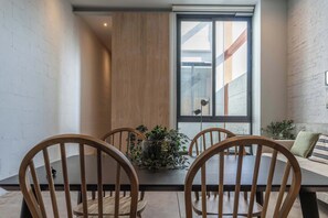 Een eetgedeelte met een houten tafel en stoelen, een kamerplant en een groot raam met uitzicht op een gebouw.