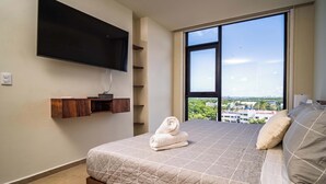 A bedroom with a large bed, a TV mounted on the wall, and a view of the city through a large window. - Comfortable with pool and full gym (Cancún)