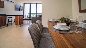 A modern dining area with a wooden table set for four, a bar area with stools, and a large window offering a view of the city. - Comfortable with pool and full gym (Cancún)